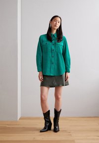 Grüne Cordhemd mit geknöpftem Vorderteil und zwei Brusttaschen, kombiniert mit einem dunkelgrünen Rock und schwarzen Stiefeletten im Western-Stil.