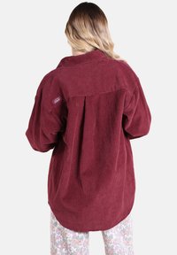 Chemise en velours côtelé bordeaux à coupe décontractée, avec un col, des manches amples et un dos plissé. Petite étiquette de marque sur l'épaule.