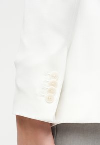 Manche de veste de costume blanche avec quatre boutons blancs assortis, portée avec un pantalon gris.