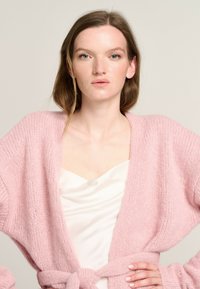 Rosa gestrickte Strickjacke mit weicher Textur, offener Vorderseite, weiten Ärmeln und einem passenden Stoffgürtel. Getragen über einem weißen Oberteil.