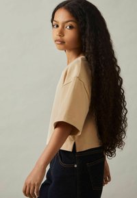 Beige kortærmet T-shirt lavet af bomuld, parret med mørkeblå denimjeans. Modellen har langt, krøllet hår.