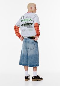 Grå grafisk t-shirt med grønt "NSX" logo, orange ternede lange ærmer og lyseblå denim culottes. Brune sko med tykke såler.