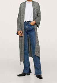 Cardigan long gris en maille avec manches larges, porté sur un t-shirt blanc et un jean bleu taille haute, assorti à des bottines noires.