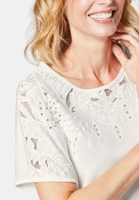 Witte top met korte mouwen, voorzien van gelaagde bloemenuitsparingen en subtiele kraalaccenten langs de halslijn, met een zachte, gladde textuur.
