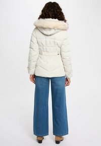 Giacca puffer crema chiaro con cappuccio in pelliccia sintetica, vita aderente e texture trapuntata. Abbinata a jeans in denim blu a gamba larga e stivaletti marroni.