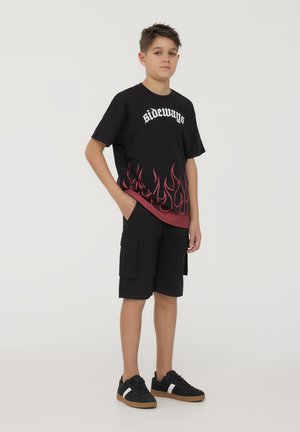 T-shirt grafiche nera con stampa di fiamme in basso e testo bianco, abbinata a pantaloni cargo neri e sneakers nere con dettagli bianchi.