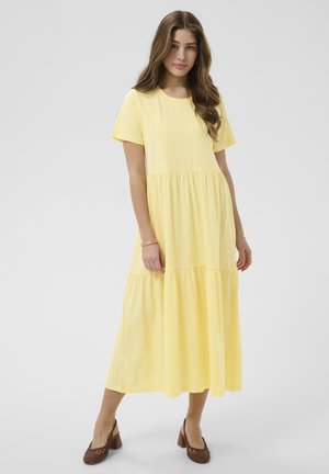 Young woman wearing a short-sleeve, yellow tiered midi dress and brown heeled woven shoes against a plain background.