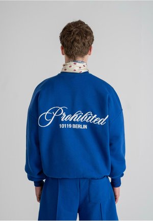 Sudadera azul con texto en cursiva blanca "Prohibido" y "10119 BERLÍN" en la parte posterior. Tejido suave, corte relajado, con puños en las muñecas.