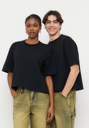 Due giovani adulti che indossano magliette nere larghe e jeans gialli sbiaditi, in piedi vicini con le mani in tasca, sorridenti con espressioni neutre.