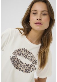 Kremo spalvos marškinėliai su dideliu leopardų rašto lūpų grafiku rudos ir juodos spalvos. Minkšta medžiaga ir laisvas kirpimas, trumpomis rankovėmis.