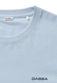 Svetlomodré bavlnené tričko s okrúhlym výstrihom, s malým logom „GABBA“ tmavomodrej farby v spodnej ľavej časti. Hladká textúra bez vzorov.