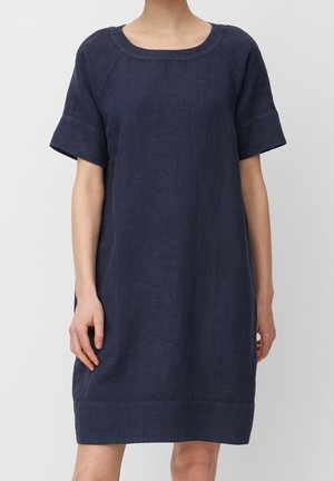 Nainen pukeutuneena väljästi istuvaan polvipituiseen tummansiniseen pellavamekkoon, jossa on lyhyet käännetyt hihat ja pyöreä pääntie.