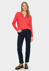 Korálový vrch s dlhým rukávom a výstrihom do V, so šikmými rozparkami, skombinovaný s tmavými denimovými džínsy a hnedými nazúvacími topánkami. Jednoduchý dizajn s voľným strihom.