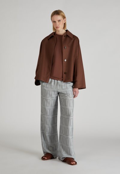 Veste oversized marron avec des boutons, assortie à un haut marron et un pantalon gris clair à motif géométrique. Sandales ouvertes marron.