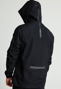 Fekete vízálló dzseki kapucnival, melynek hátsó részén függőleges cipzár és az oldalán fehér részletekkel ellátott cipzáras zseb található.