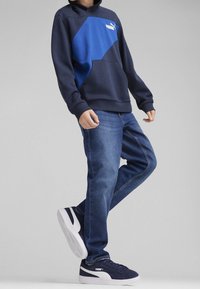 Felpa blu navy con un design geometrico blu, tasca a marsupio, abbinata a jeans di denim blu e sneakers navy con dettagli bianchi.