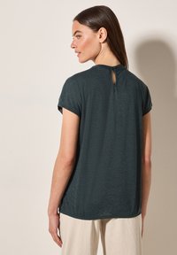 Tmavě modrá krátká rukávová halenka se uvolněným střihem, s kulatým výstřihem a detaily ve tvaru klíčové dírky na zadní straně. Hladká textilie.