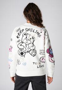 Weißes Sweatshirt mit verspielten Grafiken, darunter ein Bär sowie verschiedene bunte Kritzeleien und Sprüche, mit gerippten Bündchen und Saum.
