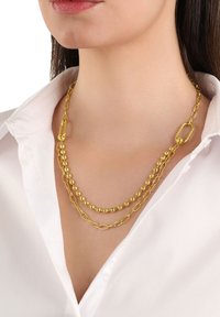 Collana oro con tre catene sovrapposte: perle rotonde, anelli allungati e un grande gancio ovale. Indossata su una camicia bianca.