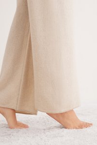 Beżowe spodnie z szerokimi nogawkami wykonane z miękkiego materiału o gładkiej fakturze, z czystym wykończeniem na krawędzi i subtelnym przeszyciem. Stopy widoczne na miękkim dywanie.