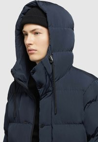 Tumesinine puffer jakk kõrge kraega ja avara kapuutsiga. Omab silmapaistvat õmblustööd, musta nööri ja nööbikinnitust. Pehme tekstuur.