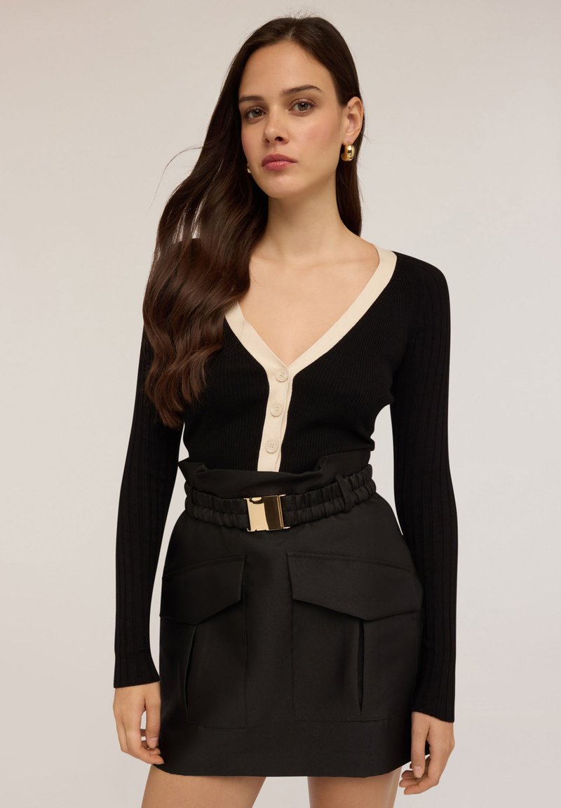 Vrouw met lang bruin haar, gekleed in een zwarte V-hals cardigan met witte rand, en een zwarte rok met grote zakken en een riem.