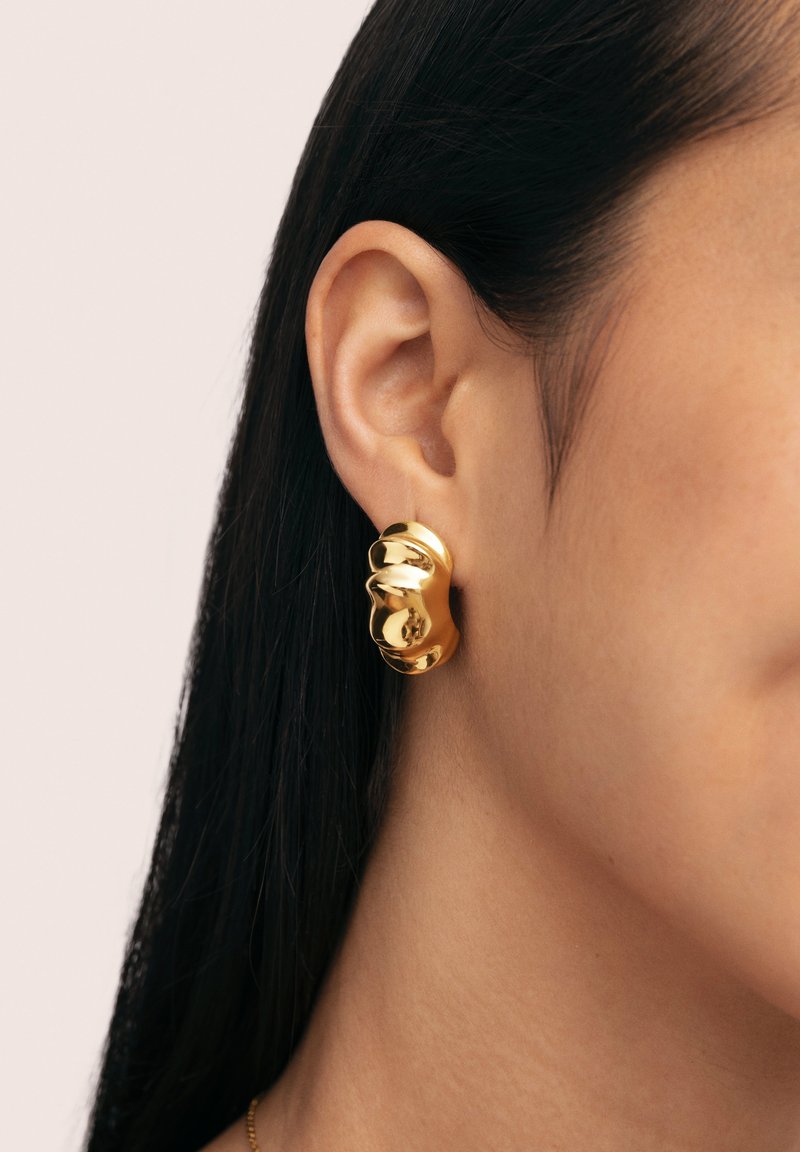 Boucles d'oreilles en or avec un design lisse et incurvé, présentant une finition métallique brillante et une surface légèrement texturée, portées à l'oreille.