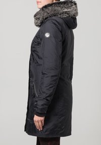 Svart vadderad jacka med huva i fuskpäls, fickor med dragkedja och ribbade muddar. Midjelång design med en strömlinjeformad silhuett.