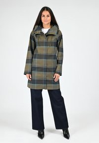 Imperméable à carreaux en olive et noir, doté d'une capuche, d'un cordon de serrage et d'une coupe droite. Porté sur un jean large sombre et des bottes à talons noires.