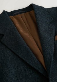 Gros plan d'un blazer en laine bleu foncé à chevrons avec une doublure intérieure marron à motifs et de larges revers à encoches.
