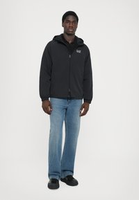 Svart zip-up jacka med huva, med elastiska muddar. Bärs över en svart skjorta, tillsammans med ljusblå utsvängda jeans och svarta skor.