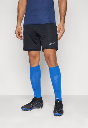 Mężczyzna ubrany w czarne sportowe spodenki Nike, niebieskie podkolanówki z wyściółką i czarne korki z niebieskimi akcentami, stojący na szarej podłodze.