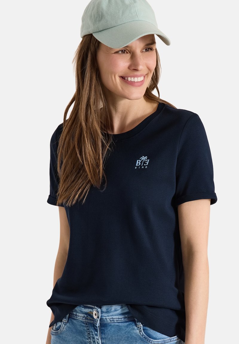 Mujer sonriente con gorra verde claro, camiseta azul marino con el logo "Be Kind" y vaqueros azules, posando frente a un fondo liso.