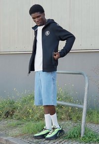 Sort zip-up hoodie med logo, hvid t-shirt, denim shorts, hvide sokker og sorte- samt neongrøn sneakers; står ved et rækværk i græs.