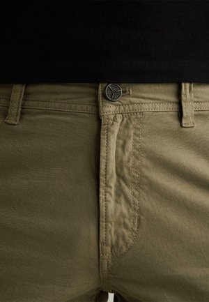 Detailný záber na pás olivovo zelených nohavíc s kovovým gombíkom a pútkami na opasok, nosené s čiernym topom zatlačeným dnu.