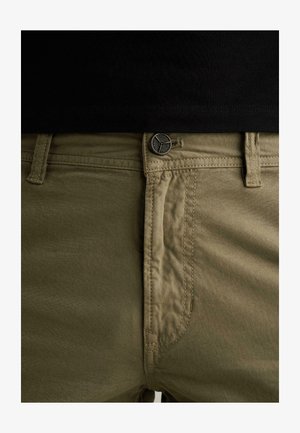 Nahaufnahme des Bunds olivgrüner Hosen mit Metallknopf und Gürtelschlaufen, getragen mit einem schwarzen, hineingesteckten Oberteil.