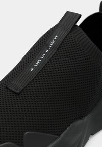 Zapatos negros sin cordones con una parte superior de malla texturizada, suela flexible y una etiqueta de tela negra con letras blancas. Diseño ligero y minimalista.