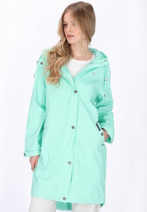 Kobieta w jasnoturkusowej, wodoodpornej kurtce z kapturem, z zatrzaskami i zamkiem błyskawicznym, pozująca z jedną ręką w kieszeni na białym tle.