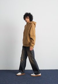 Brown hoodie with a front pocket and drawstring, paired with distressed wide-leg jeans. White sneakers with black stripes. Plain background.