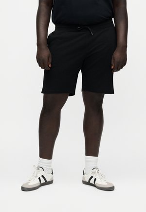 Personne portant un short noir à cordon, des chaussettes blanches montantes et des baskets blanches avec des rayures noires, debout devant un fond uni.