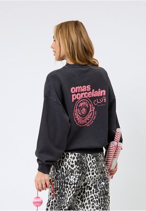 Svart genser med rosa grafisk tekst "omas porcelain CLUB" og blomstermotiv på ryggen. Har oversized passform og ribbestrikkede mansjetter.