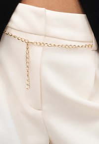 Κοντινό πλάνο σε λευκό παντελόνι με ψηλή μέση, με χρυσή ζώνη αλυσίδα και ορατό μαύρο πάνω μέρος.