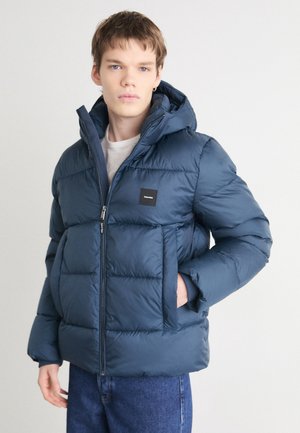 Giacca a mantello blu navy con cappuccio, chiusura con zip, polsini elastici e una patch logo quadrata sul petto. Design testurizzato e isolato.