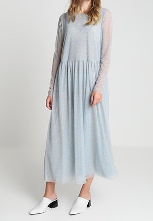 Frau trägt ein hellblaues Midikleid mit langen, transparenten Ärmeln und weiße flache Slipper, steht vor einem einfarbigen Hintergrund.