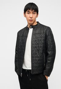 Veste matelassée noire en cuir avec un col haut, arborant un motif de logo répétitif, une fermeture éclair frontale et des poches latérales.