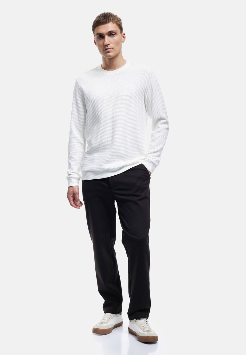 Camicia bianca a maniche lunghe con texture, abbinata a pantaloni neri e sneakers bianche con dettagli marroni. Il modello sta in piedi con le mani rilassate ai lati.