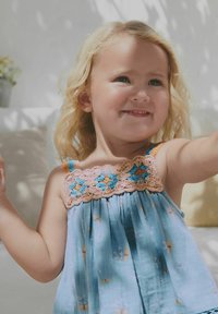 Uśmiechnięta mała dziewczynka z blond kręconymi włosami, ubrana w niebieską sukienkę w kwiaty z szydełkowymi detalami, w pomieszczeniu przy łagodnym, naturalnym świetle.