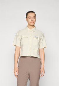 Camicetta corta a maniche corte in tessuto beige chiaro, con chiusura a bottoni, due tasche frontali e un piccolo patch con il logo sul petto.