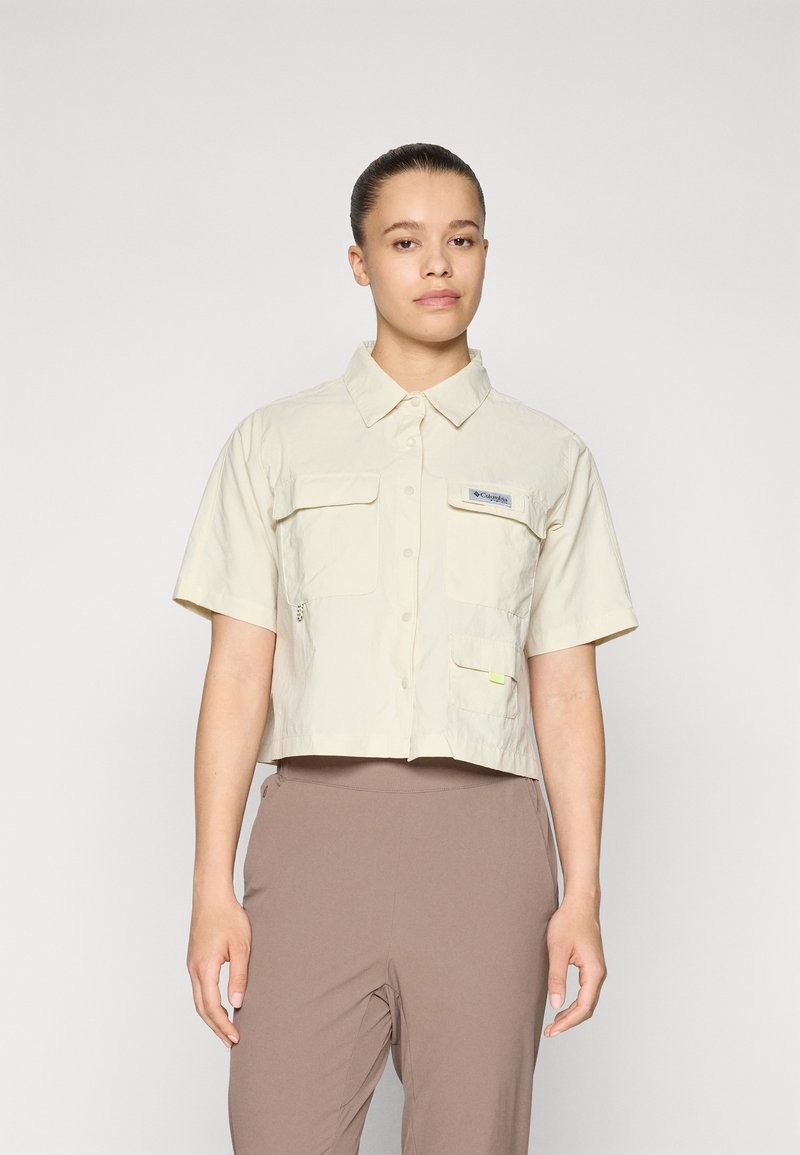Camicetta corta a maniche corte in tessuto beige chiaro, con chiusura a bottoni, due tasche frontali e un piccolo patch con il logo sul petto.