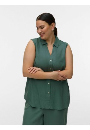 Femme aux cheveux foncés attachés, portant une chemise verte sans manches boutonnée et un pantalon assorti, souriante et les bras croisés devant un fond uni.